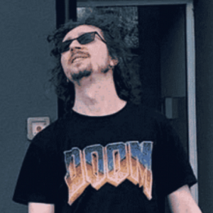 Osoba w okularach przeciwsłonecznych i koszulce z napisem "DOOM" przed budynkiem.