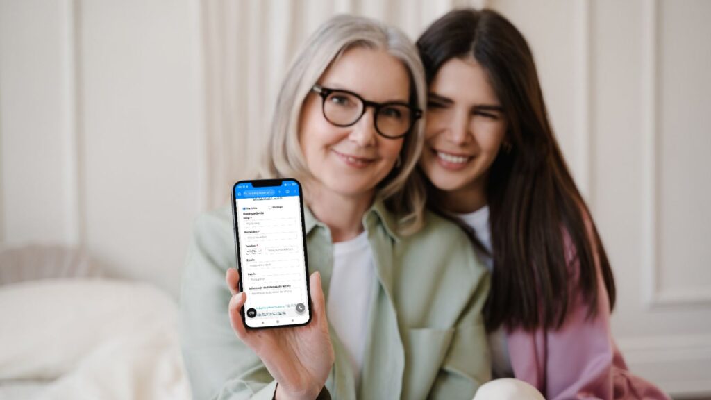 Dwie kobiety siedzą razem, jedna trzyma telefon z wypełnionym formularzem na ekranie.