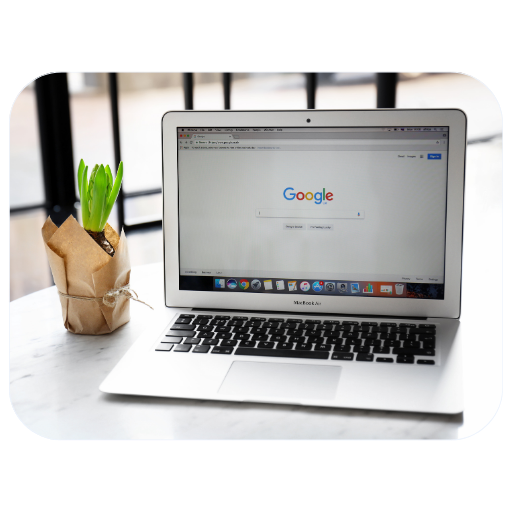 Laptop z otwartą stroną Google na stole, obok zielona roślina w papierze.