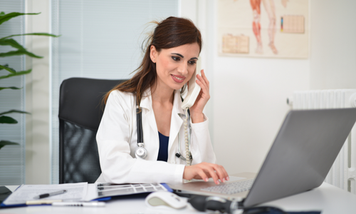 Osoba w białym fartuchu przy biurku, używa telefonu i laptopa, w tle plakat anatomiczny.
