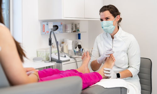 Osoba w maseczce wykonuje zabieg pielęgnacyjny na stopach w gabinecie medycznym.