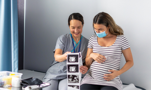 Kobieta w ciąży z maseczką ogląda z pracownikiem medycznym zdjęcia z ultrasonografii.