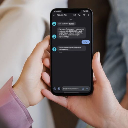 Osoba trzyma smartfon z wiadomościami o wizytach i kodzie SMS.